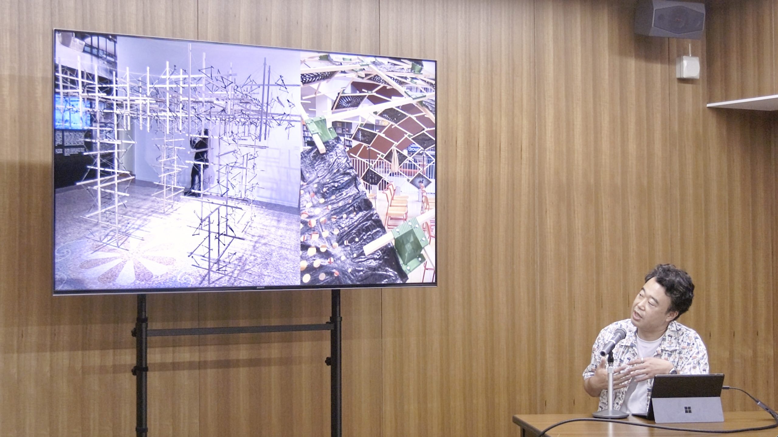 発表を行う井上准教授の写真。左側にはスクリーンがあり、発表スライド(学生作品ページ)が映し出されている。